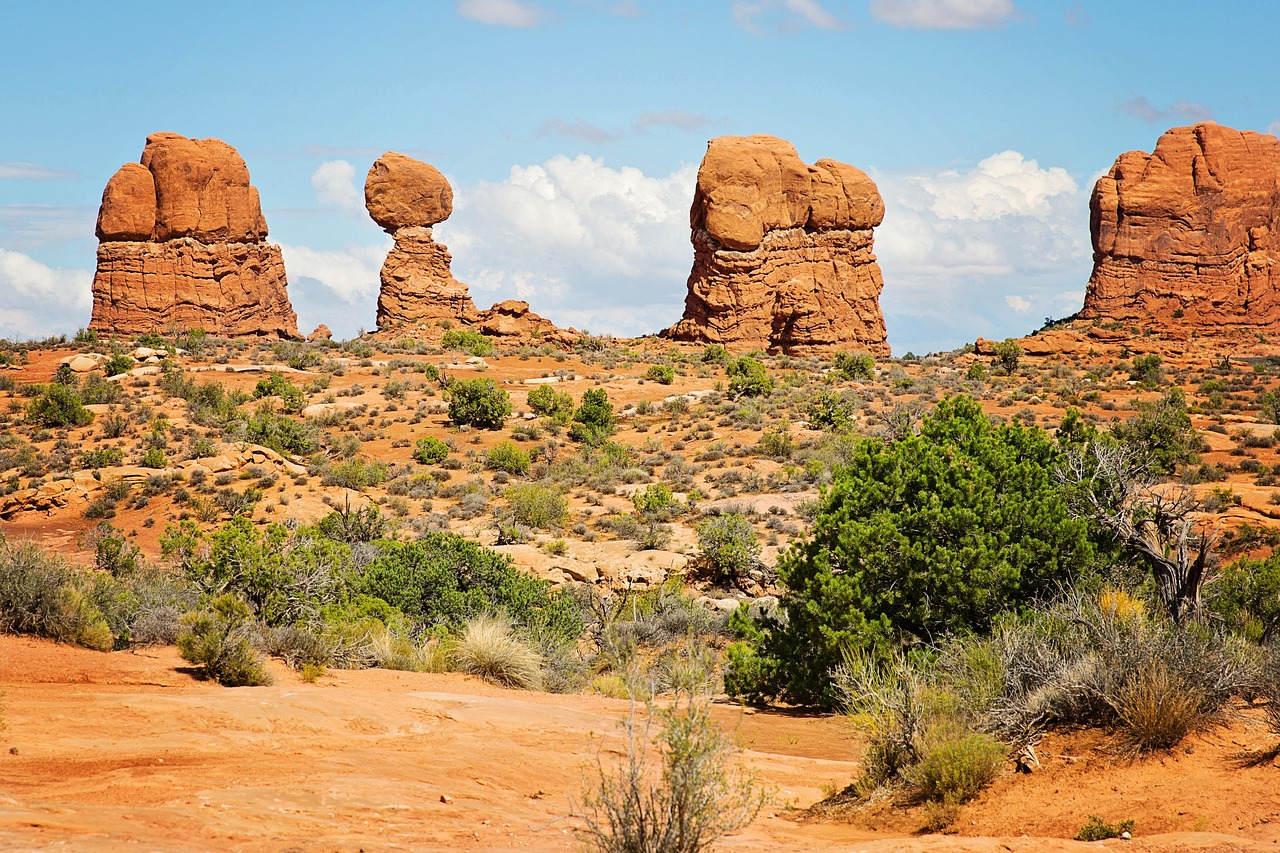Red sandstone rock formations rising from the desert near Moab, Utah, with balanced rock-like spires, desert shrubs, and a clear blue sky.