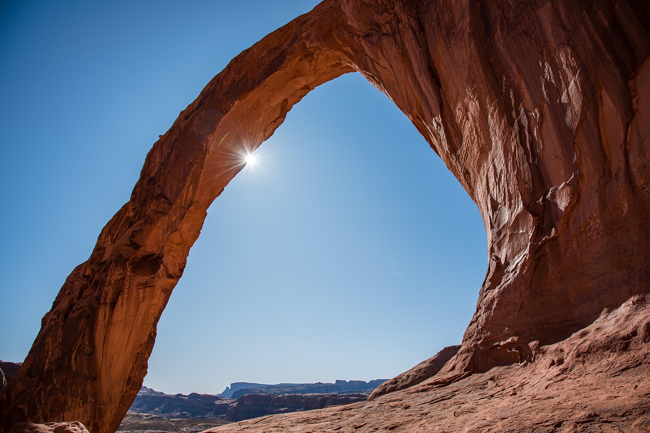 Corona Arch, a massive freestanding sandstone arch along Potash Road near Moab, Utah, with blue desert sky and red rock cliffs.