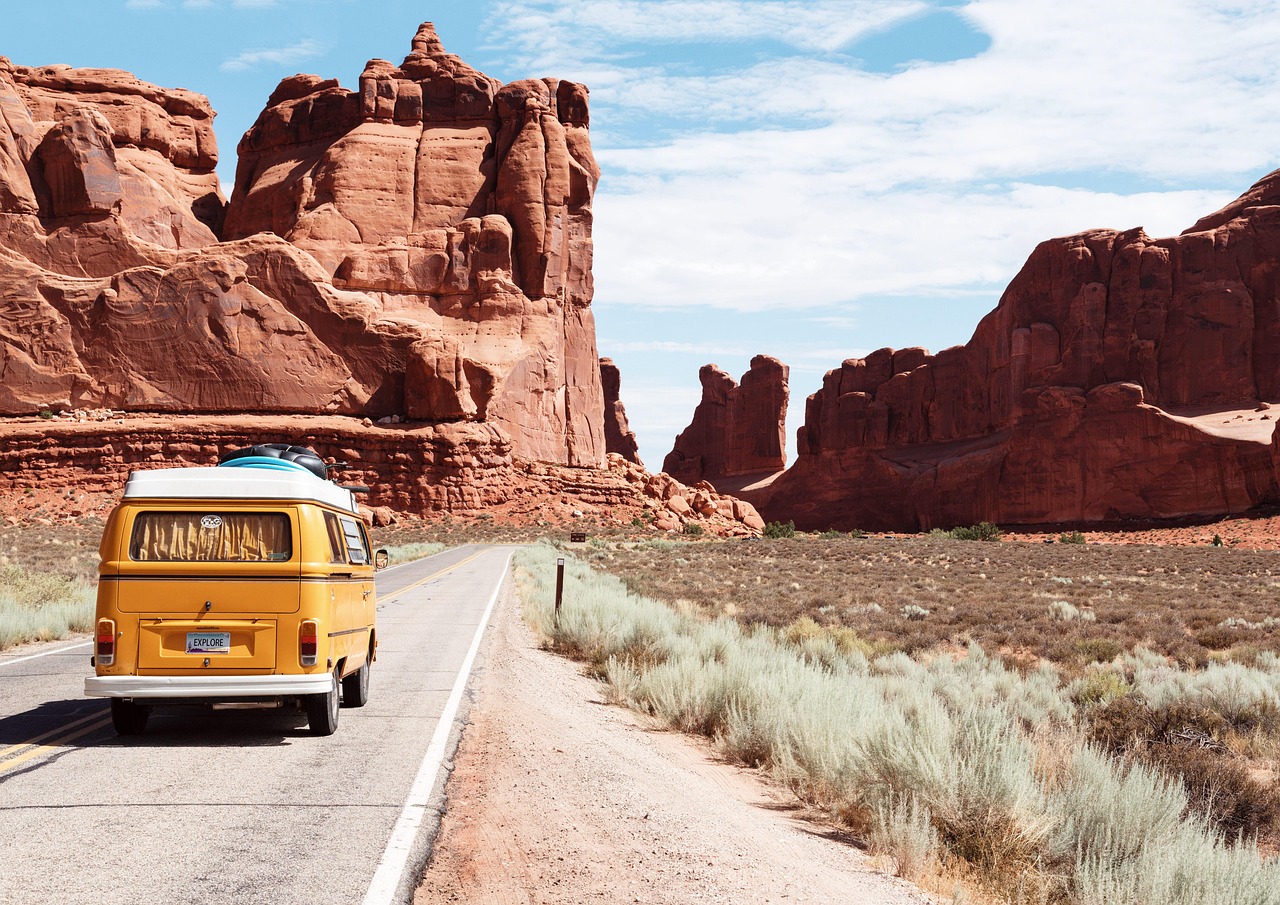 Yellow van driving on a desert highway toward towering red sandstone cliffs near Moab, Utah, under a bright blue sky.
